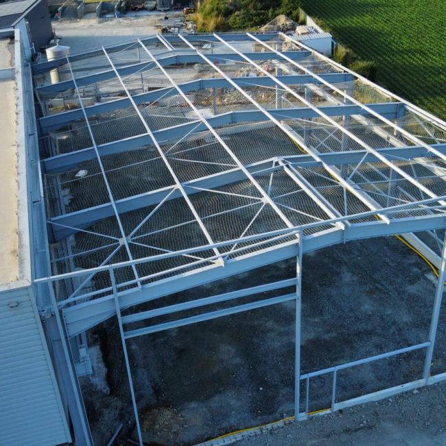 Vue aérienne d'un chantier de construction métallique montrant la structure d'un bâtiment en cours d'assemblage à côté d'un champ agricole